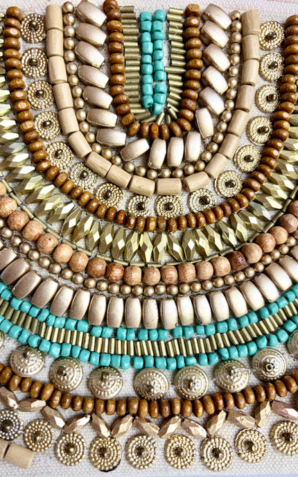 A close-up of layered bohemian beaded jewelry strands arranged in concentric semicircles, featuring natural wood beads, metallic gold and silver beads, and turquoise accents on a white textured fabric background.
