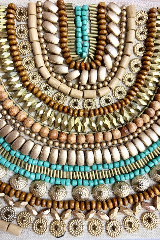 A close-up of layered bohemian beaded jewelry strands arranged in concentric semicircles, featuring natural wood beads, metallic gold and silver beads, and turquoise accents on a white textured fabric background.