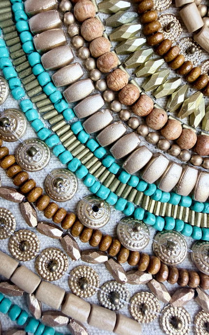 A close-up flat lay of assorted beaded strands and decorative jewelry components arranged in curved rows. The visible materials include turquoise-blue beads, cream and tan wooden beads, metallic gold and silver disc and faceted beads, and some larger round beige beads, all on a light neutral fabric background.
