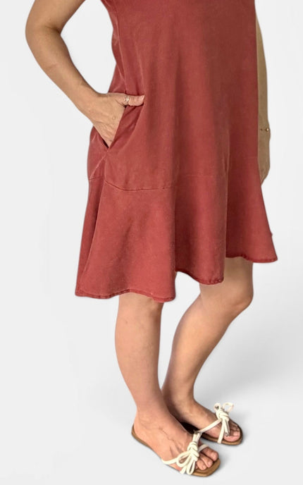 A woman modeling a sleeveless, dusty rose-colored tunic dress with side pockets, paired with white flat sandals, against a plain light gray background.