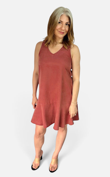 A woman modeling a sleeveless, V-neck, tiered mini dress in a dusty rose or terracotta color, paired with black flat sandals, against a light gray studio background.
