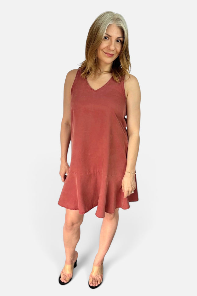 A woman modeling a sleeveless, V-neck, tiered mini dress in a dusty rose or terracotta color, paired with black flat sandals, against a light gray studio background.
