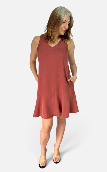 A woman modeling a sleeveless, V-neck, flared mini dress in a dusty rose or terracotta color, standing against a light gray studio background.