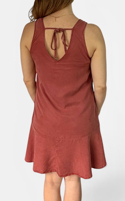 A woman shown from the back wearing a sleeveless rust-colored dress with a tie-back neckline and a flared hem, photographed against a plain light background.