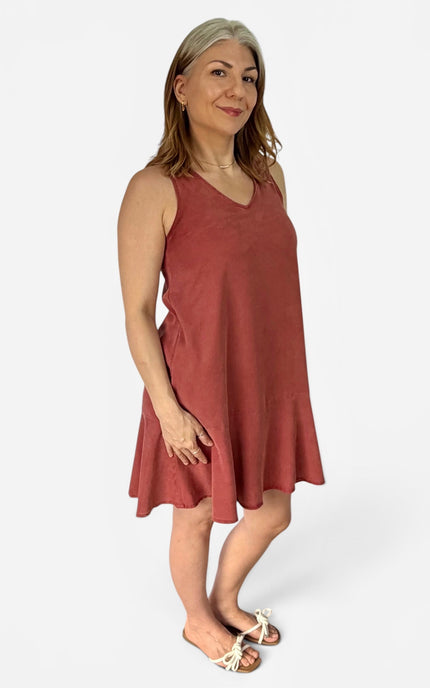 A woman modeling a sleeveless, V-neck, flared A-line dress in a dusty rose or terracotta color, paired with white strappy flat sandals, against a plain light gray studio background.