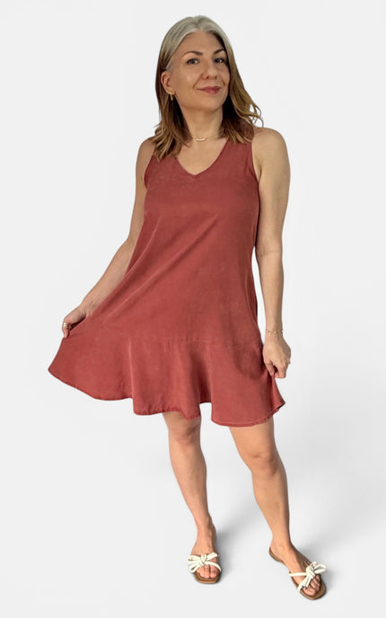 A woman modeling a sleeveless, V-neck, flowy mini dress in a dusty rose or terracotta color, paired with white slide sandals, against a plain light gray studio background.