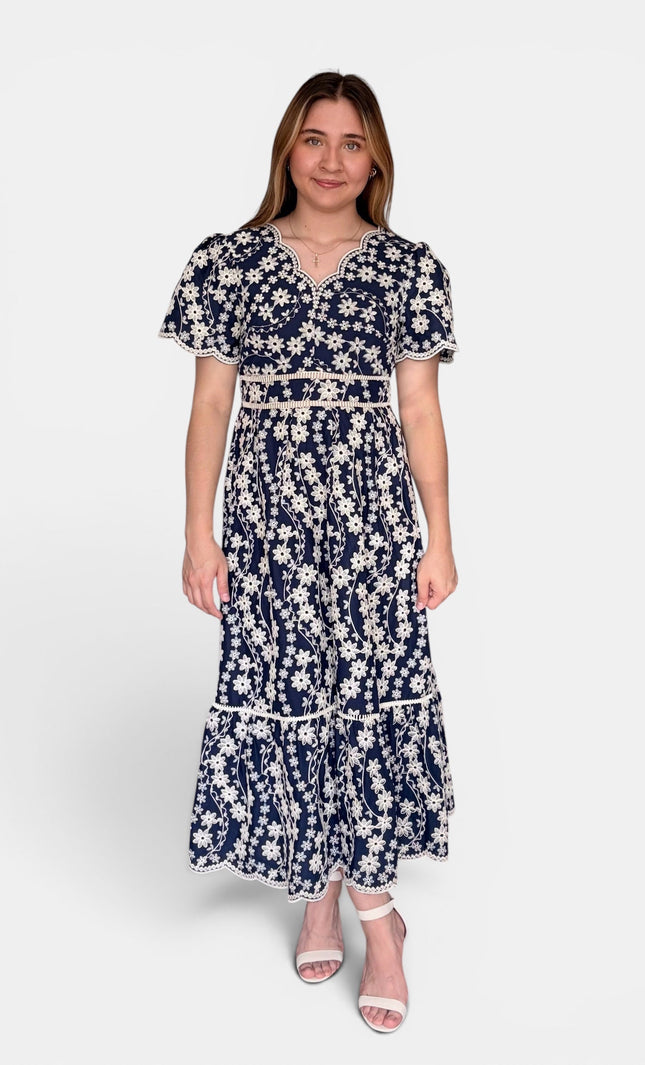 Navy blue maxi dress with a white floral embroidered pattern, featuring short flutter sleeves, a split neckline, and a tiered skirt, set against a plain light gray background.