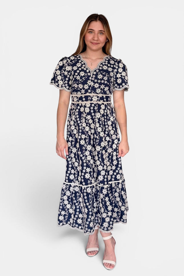 Navy blue maxi dress with a white floral embroidered pattern, featuring short flutter sleeves, a split neckline, and a tiered skirt, set against a plain light gray background.