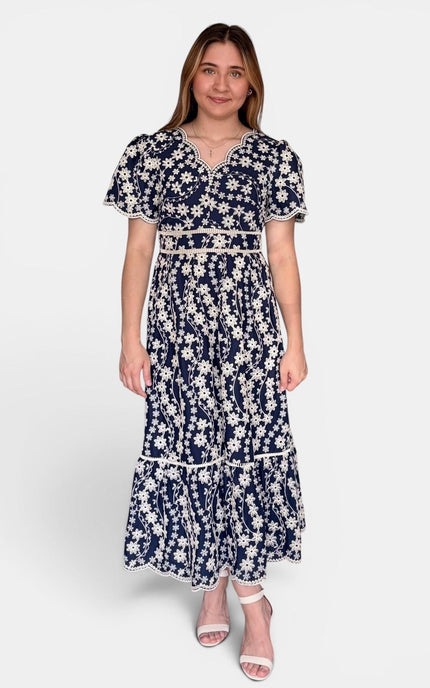 A woman modeling a navy blue maxi dress with a white floral print, featuring short puff sleeves, a V-neckline, and a matching fabric belt at the waist, set against a plain light gray background.