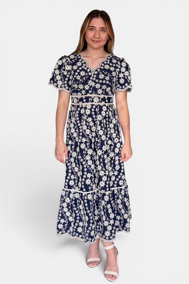 A woman modeling a navy blue maxi dress with a white floral print, featuring short puff sleeves, a V-neckline, and a matching fabric belt at the waist, set against a plain light gray background.