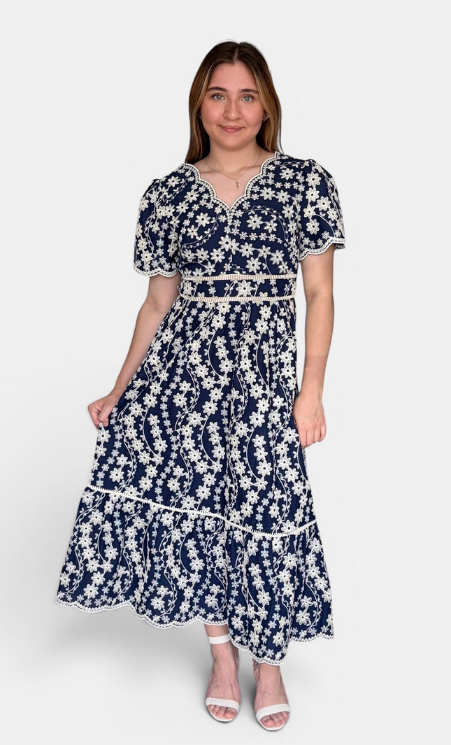 Navy blue midi dress with a white floral embroidered print, featuring short puff sleeves, a V-neckline, and a tiered skirt, set against a plain light gray background.