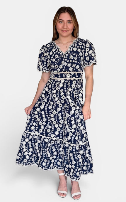 Navy blue midi dress with a white floral embroidered print, featuring short puff sleeves, a V-neckline, and a tiered skirt, set against a plain light gray background.