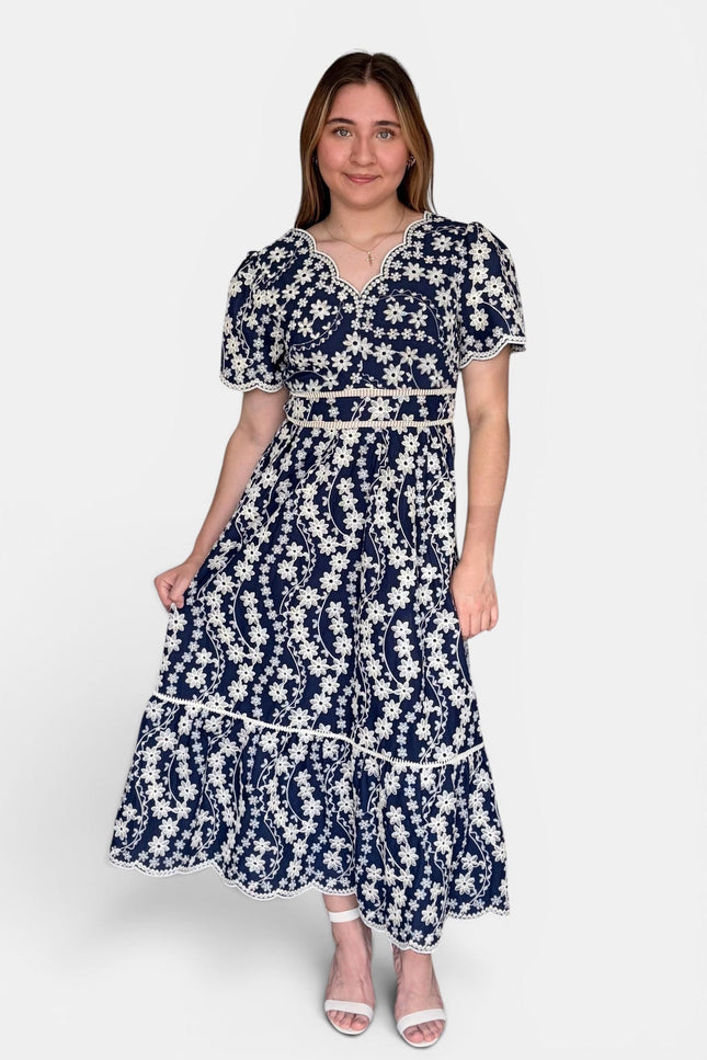 Navy blue midi dress with a white floral embroidered print, featuring short puff sleeves, a V-neckline, and a tiered skirt, set against a plain light gray background.
