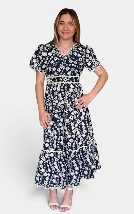 Navy blue maxi dress with a white floral print, featuring short flutter sleeves, a V-neckline, and a tiered skirt, set against a plain light gray background.