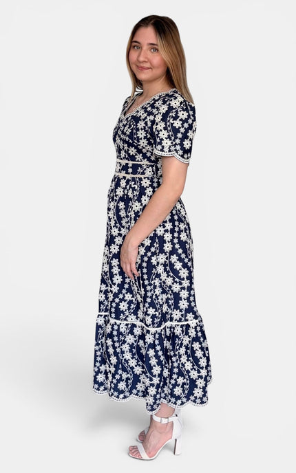 Navy blue midi dress with a white floral pattern, featuring short sleeves, a V-neckline, and a tiered skirt, paired with white high-heeled sandals against a light gray studio background.