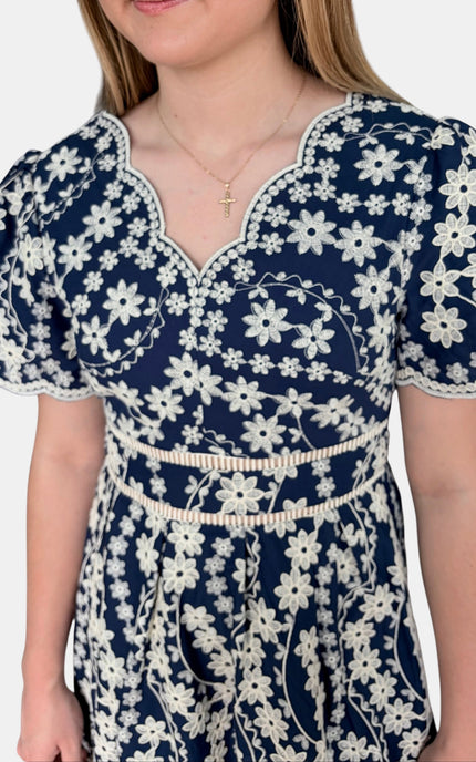 A close-up of a woman wearing a navy blue dress with a white floral pattern, featuring a scalloped V-neckline, short puff sleeves, and a matching waist tie.