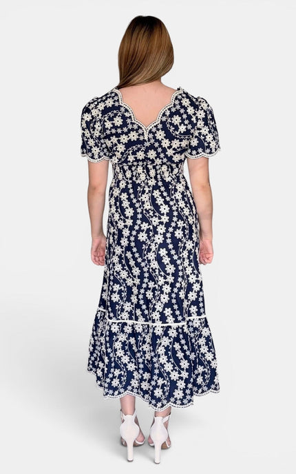A woman viewed from the back wearing a navy blue midi dress with a white floral embroidery, short puff sleeves, and a ruffled hem, paired with white high heels against a light gray studio background.