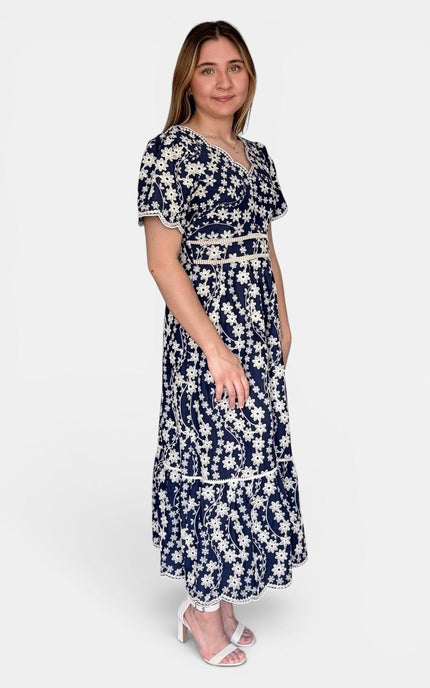 A woman modeling a navy blue maxi dress with a white floral pattern, featuring short flutter sleeves, a V-neckline, and a matching fabric belt with a decorative center detail, set against a plain light gray background.