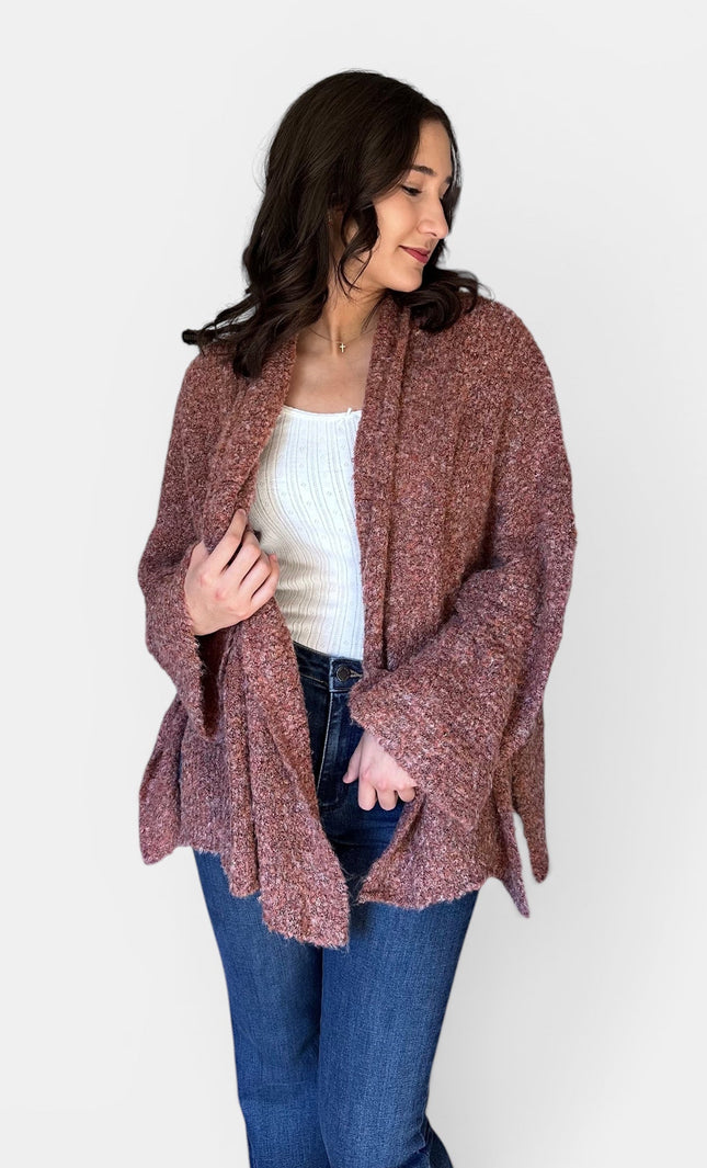 A woman wearing a chunky, open-front knit cardigan in a heathered dusty rose-brown color, styled with a white ribbed tank top and blue jeans against a plain light gray background.