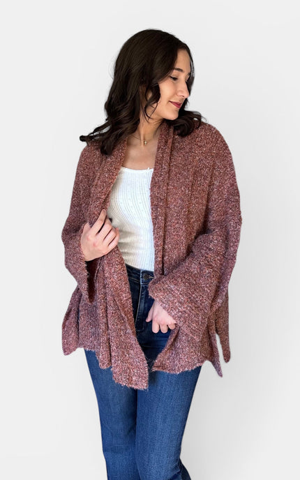 A woman wearing a chunky, open-front knit cardigan in a heathered dusty rose-brown color, styled with a white ribbed tank top and blue jeans against a plain light gray background.