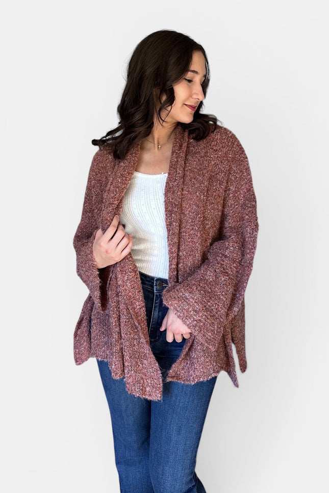 A woman wearing a chunky, open-front knit cardigan in a heathered dusty rose-brown color, styled with a white ribbed tank top and blue jeans against a plain light gray background.