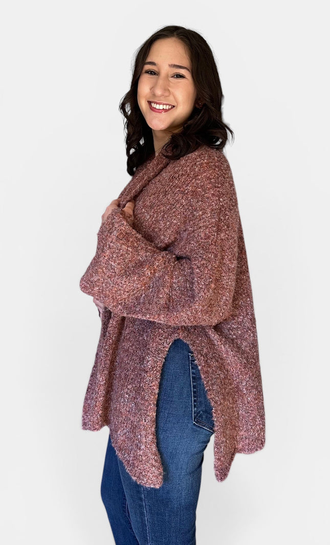 A woman modeling a loose, long-sleeve knit poncho or cardigan in a heathered dusty rose and burgundy color, paired with blue jeans, against a plain light gray background.