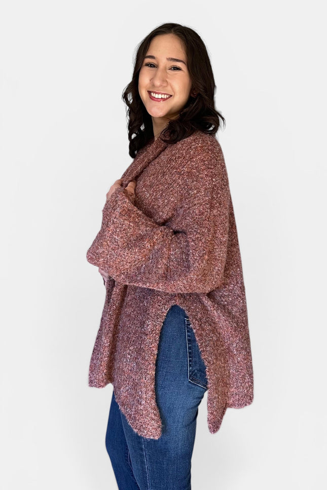 A woman modeling a loose, long-sleeve knit poncho or cardigan in a heathered dusty rose and burgundy color, paired with blue jeans, against a plain light gray background.