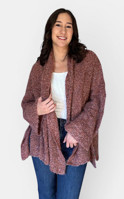 A woman modeling a chunky, open-front cardigan in a heathered dusty rose-brown color, styled with a white ribbed tank top and blue jeans against a plain white background.