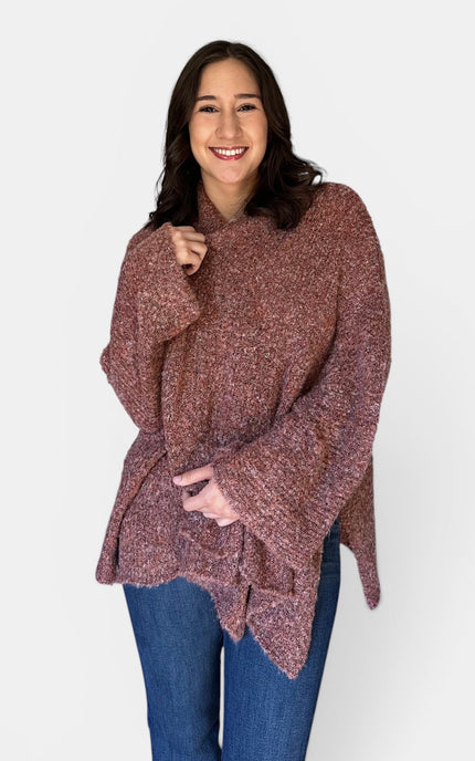 A woman wearing a chunky, fuzzy knit poncho sweater in a heathered dusty rose-brown color, paired with blue jeans, against a plain light gray background.