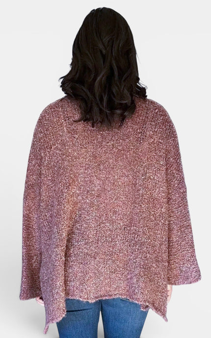 A woman viewed from behind wearing a loose, textured knit poncho sweater in a heathered dusty rose and purple color, paired with blue jeans, against a plain light gray background.