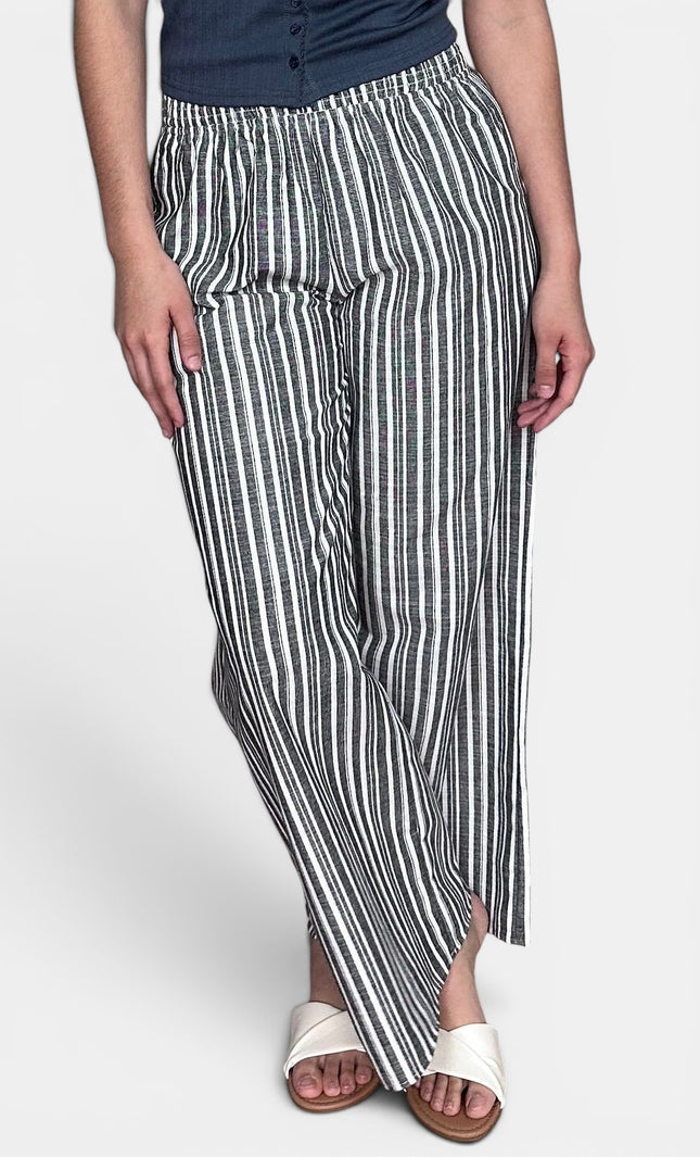 A person wearing loose, wide-leg striped pants with vertical black and off-white stripes, paired with a dark top and white slide sandals, against a light gray studio background.