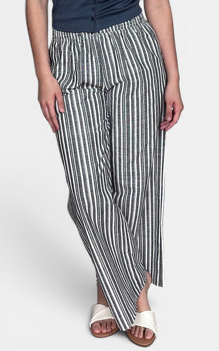 A person wearing loose, wide-leg striped pants with vertical black and off-white stripes, paired with a dark top and white slide sandals, against a light gray studio background.
