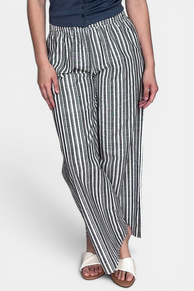 A person wearing loose, wide-leg striped pants with vertical black and off-white stripes, paired with a dark top and white slide sandals, against a light gray studio background.