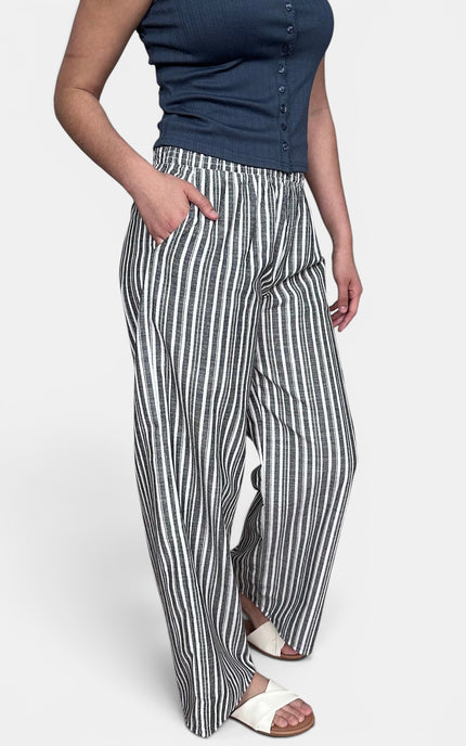 A model wearing high-waisted, wide-leg trousers with vertical black and white stripes, paired with a navy blue sleeveless top and white slide sandals, against a light gray studio background.