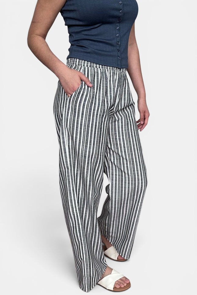 A model wearing high-waisted, wide-leg trousers with vertical black and white stripes, paired with a navy blue sleeveless top and white slide sandals, against a light gray studio background.