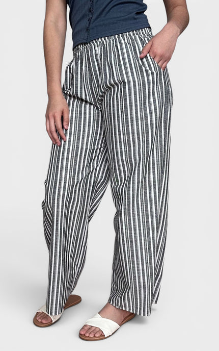 A person wearing loose, wide-leg striped pants with vertical black and white stripes, paired with a navy top and white slide sandals, against a light gray studio background.