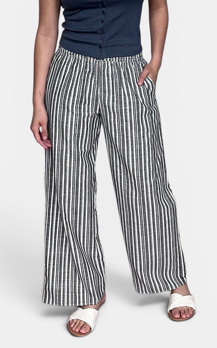 A model wearing a pair of wide-leg pants with vertical black and white stripes, paired with a navy blue top and white slide sandals, shown from the waist down against a light gray background.
