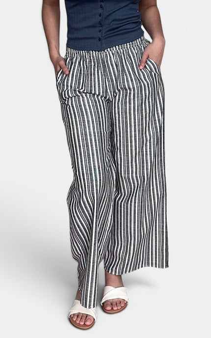 A person wearing loose, wide-leg striped pants with a navy top and white slide sandals, shown from the waist down against a light gray studio background.