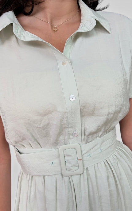 A close-up of a woman wearing a light sage green short-sleeve shirt dress with a matching fabric belt and square buckle.