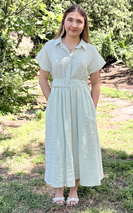 A woman modeling a light sage green tence midi shirt dress with pockets and short sleeves, a buttoned collar, and a matching fabric belt with a square buckle, paired with white slide sandals against a plain light gray studio background.