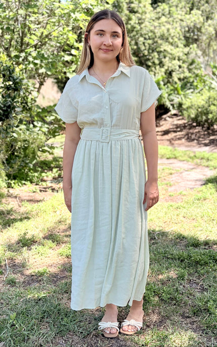 A woman modeling a light sage green tencel midi shirt dress with pockets and short sleeves, a buttoned collar, and a matching fabric belt with a square buckle, paired with white slide sandals against a plain light gray studio background.