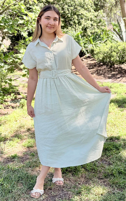 A woman modeling a light sage green midi shirt dress with short sleeves, a buttoned collar, and a matching fabric belt with a square buckle, paired with white slide sandals against a plain light gray studio background.