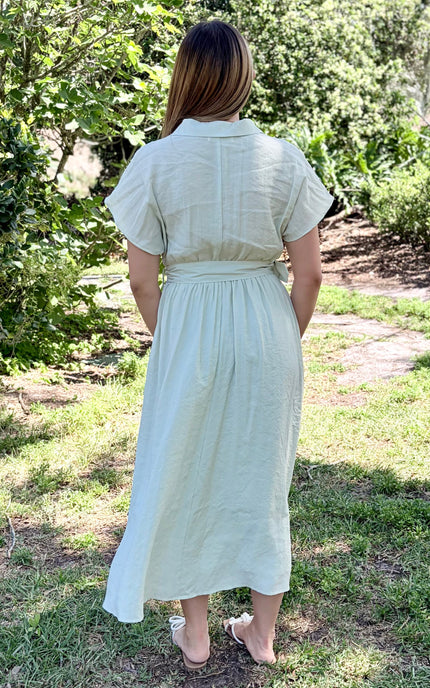 A woman modeling the back of a tencel light sage green midi shirt dress with pockets and short sleeves, a buttoned collar, and a matching fabric belt with a square buckle, paired with white slide sandals against a plain light gray studio background.