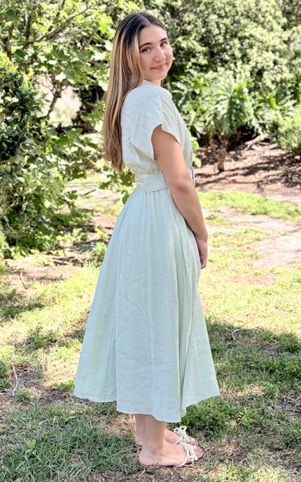 A side view of a light sage green tencel midi shirt dress with pockets and short sleeves, a buttoned collar, and a matching fabric belt with a square buckle, paired with white slide sandals against a plain light gray studio background.
