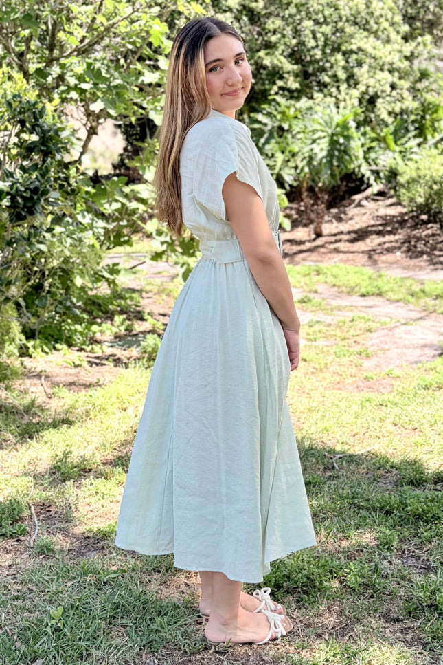 A side view of a light sage green tencel midi shirt dress with pockets and short sleeves, a buttoned collar, and a matching fabric belt with a square buckle, paired with white slide sandals against a plain light gray studio background.