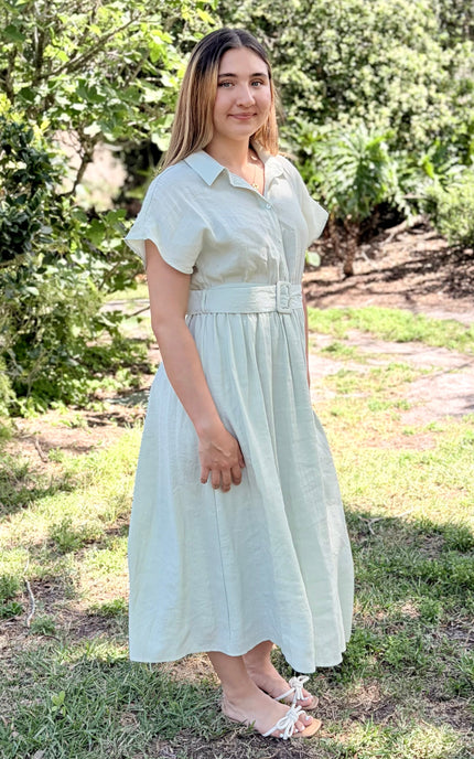 A side view of a light sage green tencel midi shirt dress with pockets and short sleeves, a buttoned collar, and a matching fabric belt with a square buckle, paired with white slide sandals against a plain light gray studio background.