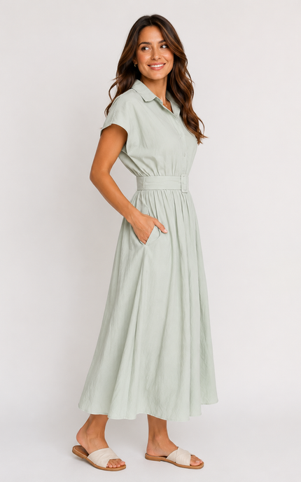 A woman modeling a light sage green midi shirt dress with pockets and short sleeves, a buttoned collar, and a matching fabric belt with a square buckle, paired with white slide sandals against a plain light gray studio background.