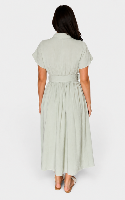 A woman viewed from the back wearing a light sage green midi shirt dress with short sleeves and a matching tie belt, paired with tan flat sandals against a plain white studio background.