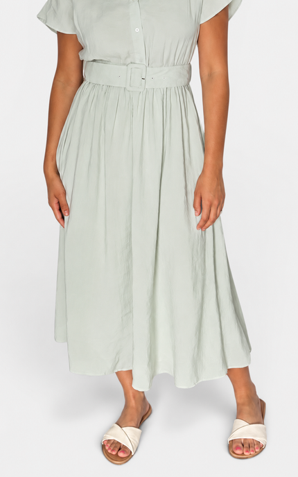 A woman wearing a light sage green midi dress with a matching waist belt and white slide sandals, shown from the chest down against a plain light gray studio background.