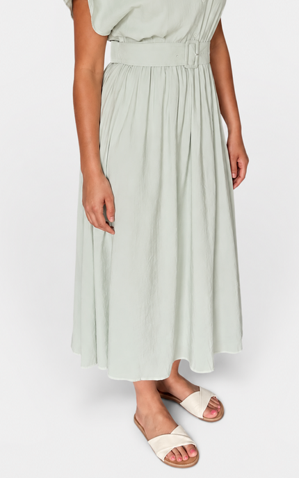 A woman wearing a light sage green midi dress with short flutter sleeves, a collared neckline, and a matching fabric tie belt at the waist, paired with cream slide sandals, against a plain light gray studio background.